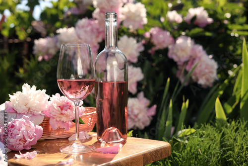 Fototapeta Naklejka Na Ścianę i Meble -  Bottle and glass of rose wine near beautiful peonies on wooden table in garden. Space for text