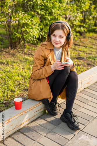 A young teenage girl sits on the street with a smartphone in her hand, headphones, listens to music. High quality photo