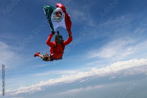 The national flag of Mexico is in the sky. A red skydiver is flying in the sky with Mexican flag.