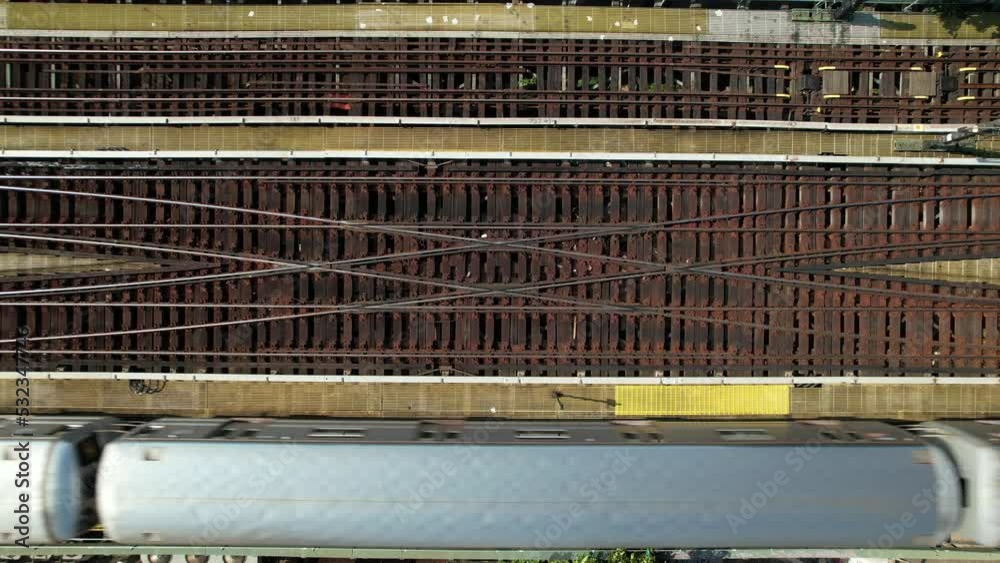 A top down view, directly above elevated train tracks as a silver subway travels from left to right of the frame on a sunny day. The drone camera is stationary.