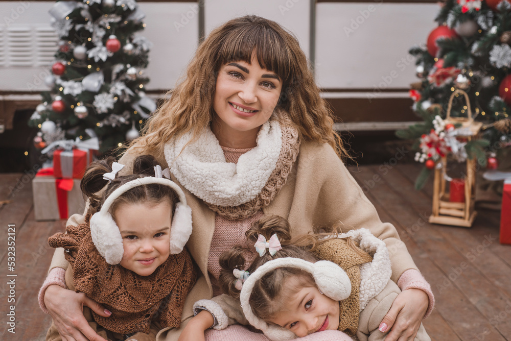 Mom and two twin daughters dressed in stylish fur coats and winter fur headphones play, hug and enjoy near the trailer in the forest with New Year's decorations. Christmas atmosphere and comfort Mom and two twin daughters dressed in stylish fur coats and winter fur headphones play, hug and enjoy near the trailer in the forest with New Year's decorations. Christmas atmosphere and comfort