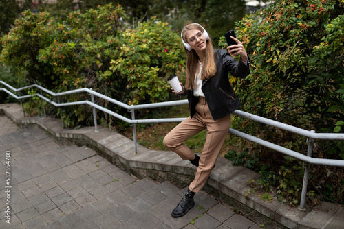 young woman in the park listening to music in headphones with a cup of coffee and taking a selfie