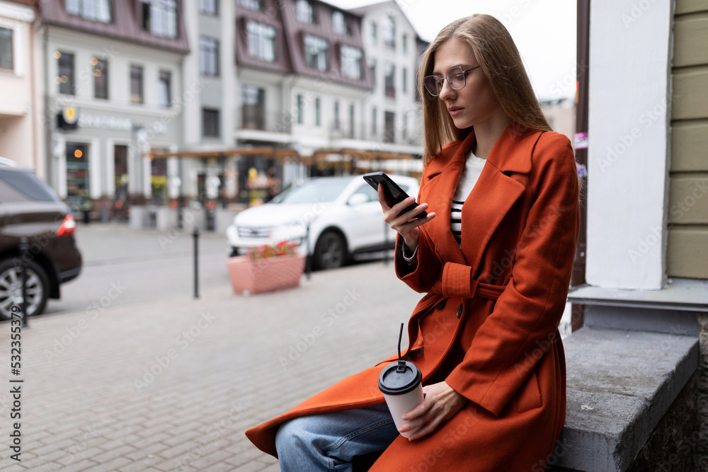 a young student with a cup of coffee looks at a mobile phone on the background of the city