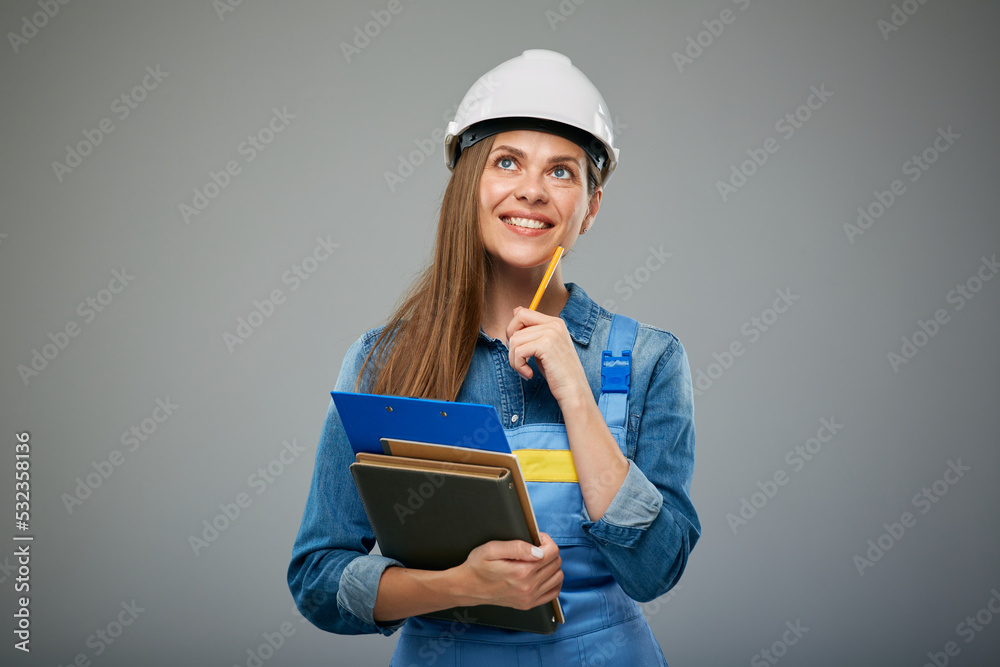 Thinking woman engineer wearing white helmet and overall holding books and clipboard. Isolated female builder portrait. Thinking woman engineer wearing white helmet and overall holding books and clipboard. Isolated female builder portrait.