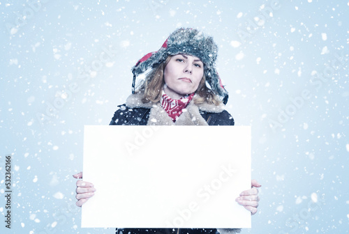 Frozen young woman in winter clothes holds an empty white poster in her hands, it is snowing on a blue background