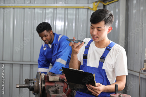 Multiethnic technicain mechanic using metal lathe machine operate polishing car disc brake with smart mobile phone and clipboard at garage. Maintenance automotive and inspecting vehicle part concept photo