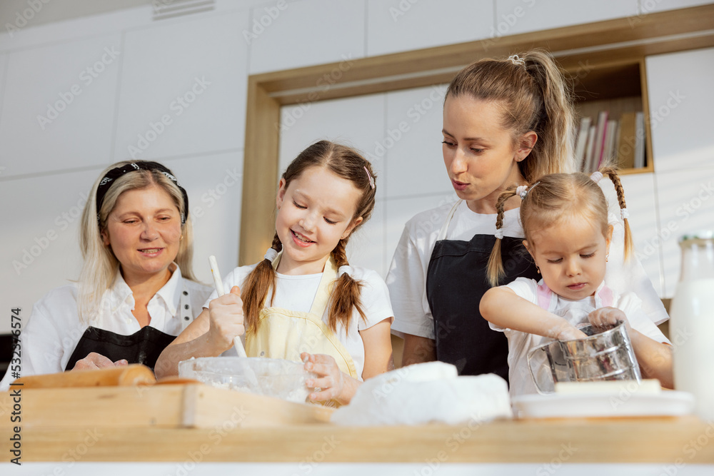 Young girl kneading dough using ingredients eggs flour milk butter. Granny managing process smiling. Mother with preschooler daughter sprinkling flour on metal sieve. Young girl kneading dough using ingredients eggs flour milk butter. Granny managing process smiling. Mother with preschooler daughter sprinkling flour on metal sieve.