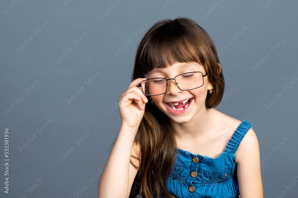 A funny positive little girl put on adult glasses on her face and laughs merrily and smiles with fallen milk teeth. Ophthalmologist and vision care. Emotional photo, happy preschool child