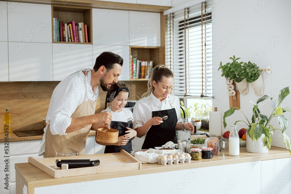 Happy family in aprons holding sieve preparing piza ingredients. Mother holding spoon taking flour pouring on sieve. Curious daughter watching process.