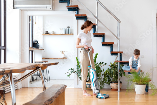 woman with cordless vacuum cleaner cleans floor, preschool child on appartment