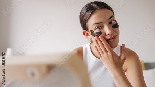 Pretty young caucasian female smoothers moisturizing patches under her eyes. Brunette hair girl sitting on bed in front of mirror. Concept good morning, beauty.