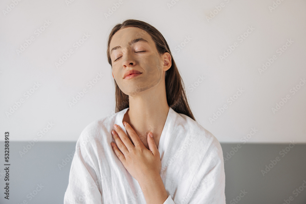 Close-up young caucasian woman with cleansing clay on face, sits on bed with closed eyes, gently touching herself. Brown hair girl enjoying beauty procedures at home. Concept beauty, tenderness. Close-up young caucasian woman with cleansing clay on face, sits on bed with closed eyes, gently touching herself. Brown hair girl enjoying beauty procedures at home. Concept beauty, tenderness.