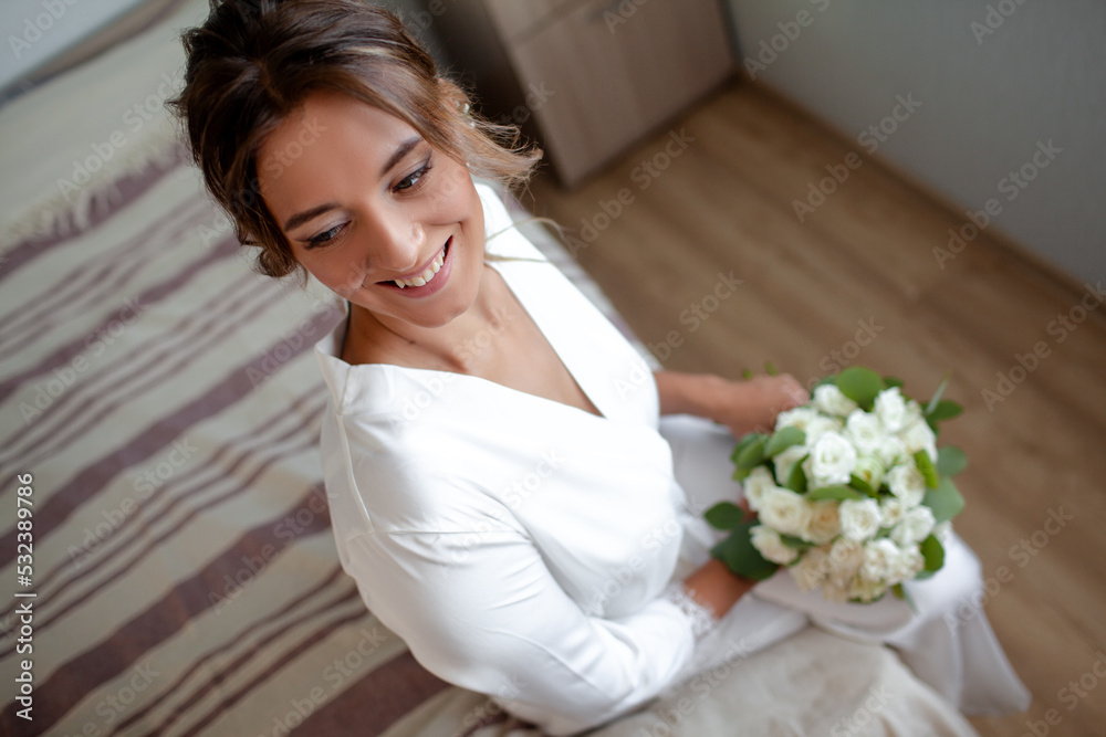 Beautiful bride dressed in a wedding white robe, holds a bride's bouquet and smiles, preparation for the wedding, brise's morning Beautiful bride dressed in a wedding white robe, holds a bride's bouquet and smiles, preparation for the wedding, brise's morning