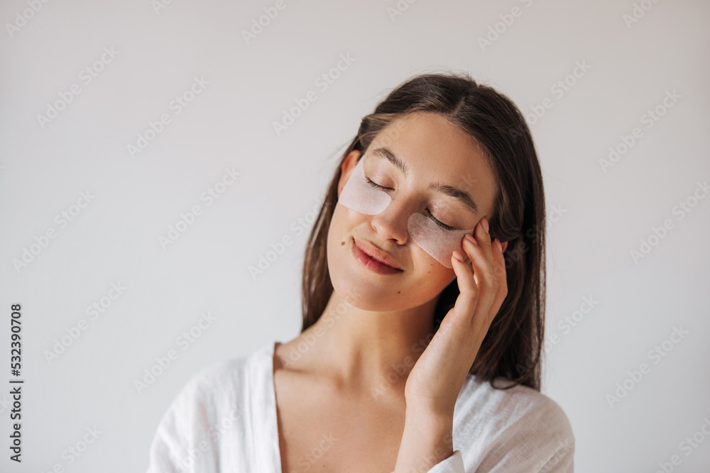 Close up tender young caucasian woman gently touching her face with closed eyes at home. Women's brown hair, wears white clothes. Skin care concept Close up tender young caucasian woman gently touching her face with closed eyes at home. Women's brown hair, wears white clothes. Skin care concept