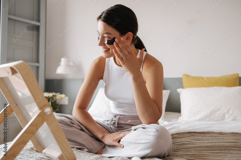 Brunette hair young caucasian girl uses products to smooth wrinkles on perfect skin. Pretty female sitting on bed and smiling with teeth. Concept positive morning, beauty.