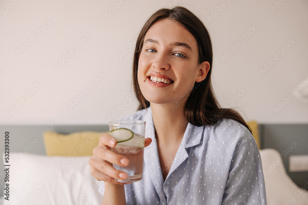 Positive young caucasian girl smiling with teeth and looking at camera. Brunette hair woman with glass of water sitting on bed. Concept enjoying weekend. Positive young caucasian girl smiling with teeth and looking at camera. Brunette hair woman with glass of water sitting on bed. Concept enjoying weekend.