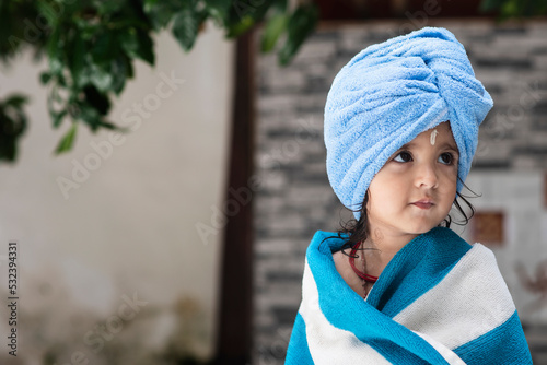 Fotografie 22 months old cute  Indian baby girl wearing blue Hair Towel Wrap Turban and body wrapped with blue and white towel, sandalwood anoint on her forehead sitting and looking side wise