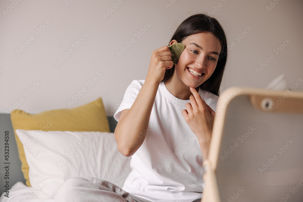Happy young caucasian girl doing facial massage with scraper with big smile at home. Brunette sits on bed in her bedroom. Skin beauty concept, positive morning. Happy young caucasian girl doing facial massage with scraper with big smile at home. Brunette sits on bed in her bedroom. Skin beauty concept, positive morning.