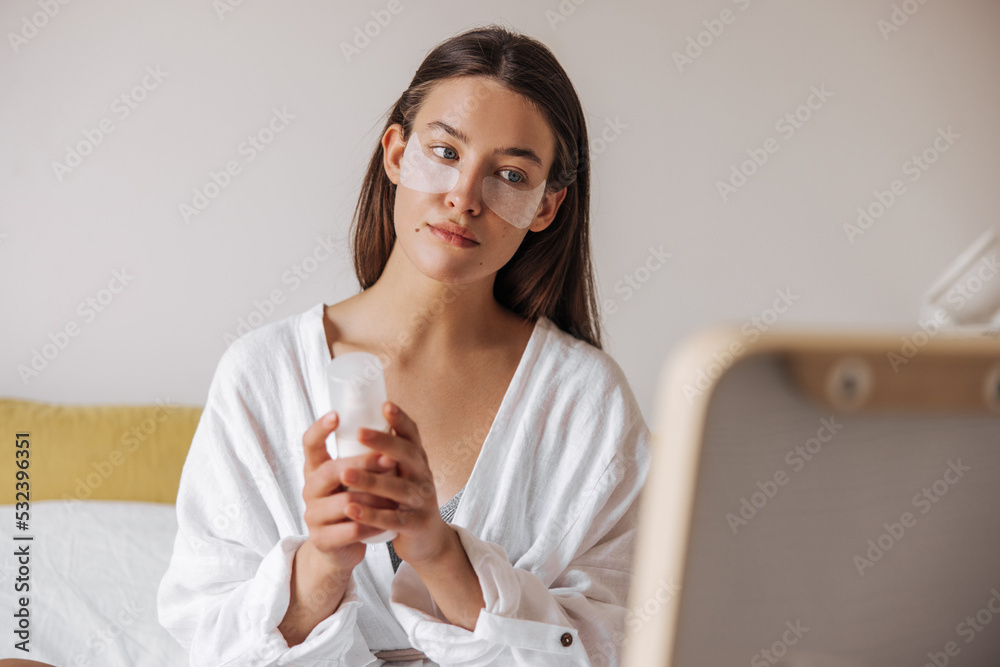 Calm young caucasian woman with eyes patches holding face cream jar at home. Brunette hair female wears white shirt in front of mirror. Concept beauty skin, care. Calm young caucasian woman with eyes patches holding face cream jar at home. Brunette hair female wears white shirt in front of mirror. Concept beauty skin, care.