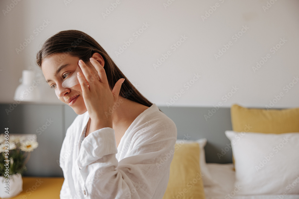 Cute young caucasian woman smoothes patches under eye sitting on bed in morning. Brunette hair girl wears white shirt with pillows and flower in background. Concept moisturizing and smoothing wrinkles Cute young caucasian woman smoothes patches under eye sitting on bed in morning. Brunette hair girl wears white shirt with pillows and flower in background. Concept moisturizing and smoothing wrinkles