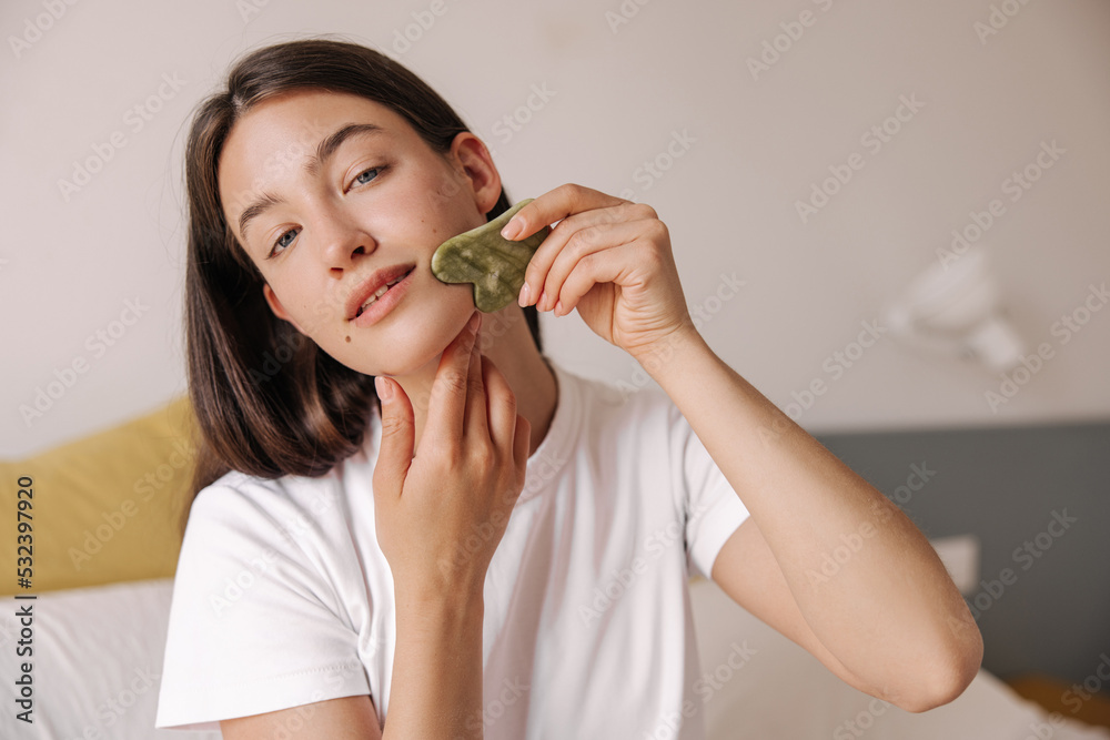 Close-up of beautiful young caucasian woman in morning smoothes wrinkles on her face with scraper. Girl brunette hair uses massager. Concept beauty, skin care. Close-up of beautiful young caucasian woman in morning smoothes wrinkles on her face with scraper. Girl brunette hair uses massager. Concept beauty, skin care.