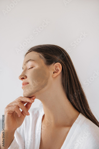 Close-up young caucasian girl gently touches her face with closed eyes sitting on bed in bedroom. Brunette hair female wears white pajamas on light background. Concept tenderness, beauty.