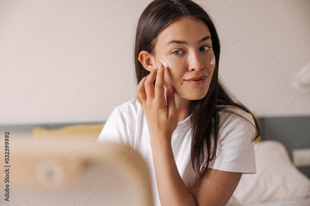 Pretty young caucasian girl applying cream on her face looking in mirror at home. Brunette hair woman wears white t-shirt. Concept beauty rituals. Pretty young caucasian girl applying cream on her face looking in mirror at home. Brunette hair woman wears white t-shirt. Concept beauty rituals.