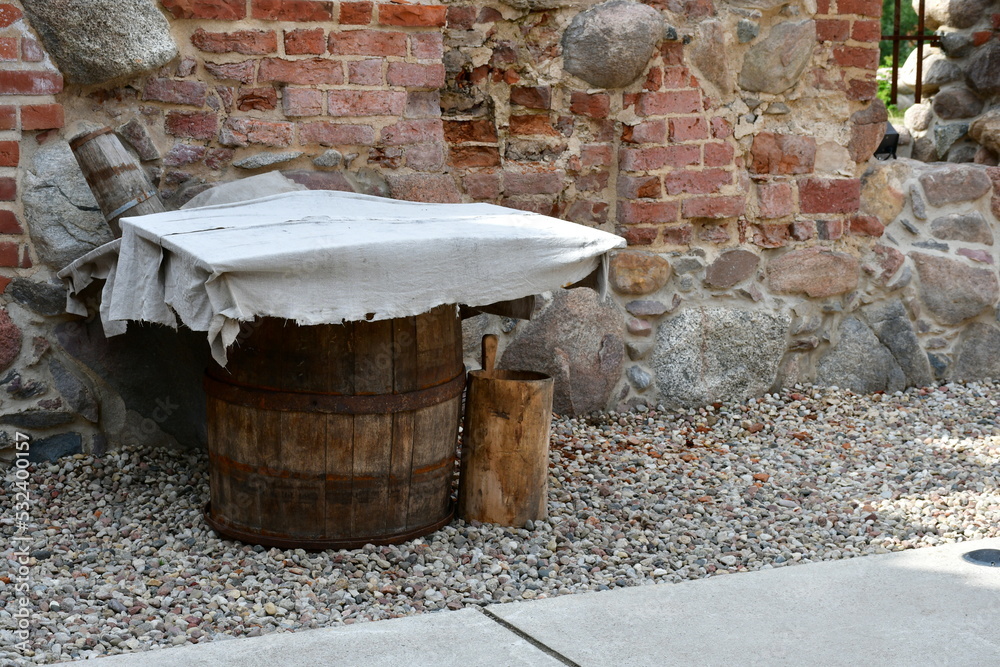 A close up on a wooden barrel made out of planks, logs, and boards standing in the middle of a gravel path covered with a white cloth standing next to a red brick castle or fence in Poland A close up on a wooden barrel made out of planks, logs, and boards standing in the middle of a gravel path covered with a white cloth standing next to a red brick castle or fence in Poland