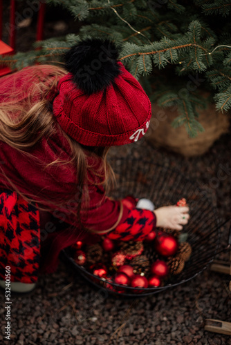 A girl in a red coat and hat decorates the Christmas tree with red balls at the Christmas tree market