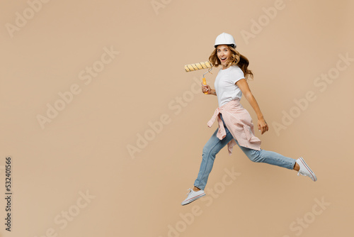 Full body young employee laborer handyman woman in white t-shirt helmet jump high hold wall paint roller isolated on plain beige background Instruments accessories renovation room Repair home concept