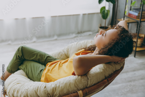 Side view young woman of African American ethnicity wear casual clothes sits in armchair hold hands behind neck take nap stay at home flat relax spend free spare time in living room indoors grey wall.