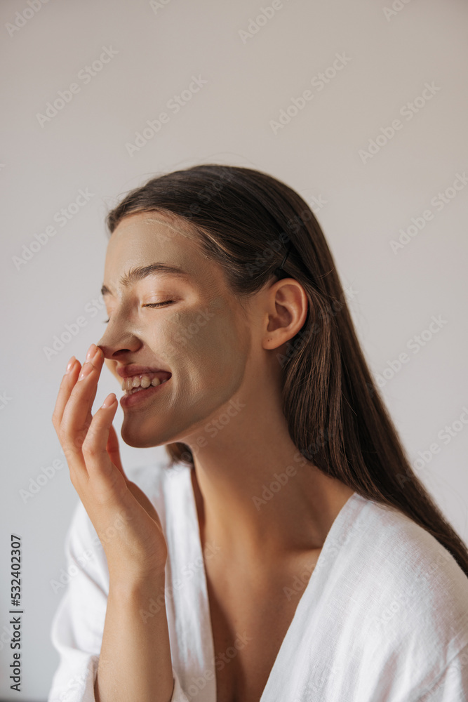 Happy young caucasian woman with skin care product on her face wide smiling teeth at home. Brunette hair female wears in pajamas enjoying morning procedures. Concept positive emotions, beauty shooting Happy young caucasian woman with skin care product on her face wide smiling teeth at home. Brunette hair female wears in pajamas enjoying morning procedures. Concept positive emotions, beauty shooting