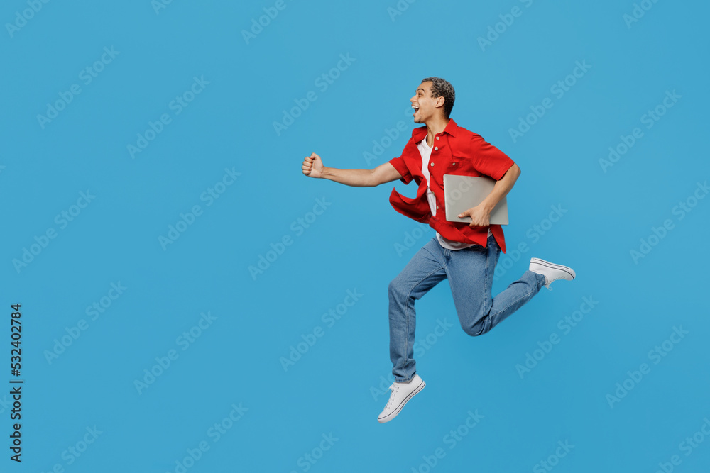 Full body side view young IT man of African American ethnicity wear red shirt hold use work on laptop pc computer jump high run isolated on plain pastel blue cyan background. People lifestyle concept.