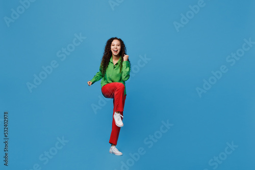 Full body young woman of African American ethnicity 20s she wear green shirt doing winner gesture celebrate clenching fists say yes isolated on plain blue background studio. People lifestyle concept.