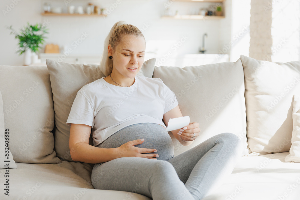Happy pregnant woman looking at ultrasound scan of baby on sofa, light kitchen background Happy pregnant woman looking at ultrasound scan of baby on sofa, light kitchen background