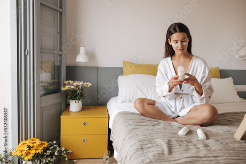 Beautiful young caucasian woman wears in casual shirt sitting on bed. Brown hair girl study composition of cream for facial skin care. Concept morning 