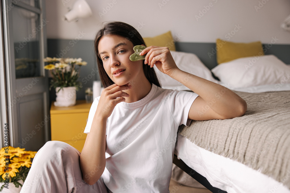 Relaxed young caucasian woman is sitting on floor near bed and takes care of her face skin by massager. Brunette hair girl wears pajamas at home. Concept beauty procedures, massage. Relaxed young caucasian woman is sitting on floor near bed and takes care of her face skin by massager. Brunette hair girl wears pajamas at home. Concept beauty procedures, massage.