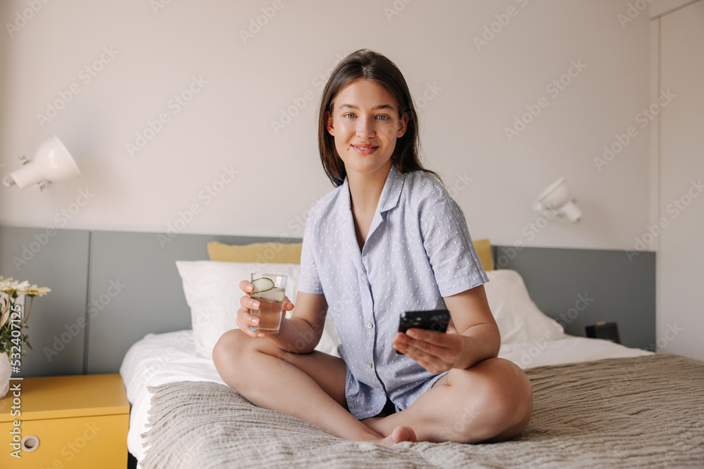 Beautiful young caucasian girl keeps glass of water and using her smartphone on bed. Brunette hair woman dressed in pajamas. Concept cozy vacation, gadgets. Beautiful young caucasian girl keeps glass of water and using her smartphone on bed. Brunette hair woman dressed in pajamas. Concept cozy vacation, gadgets.