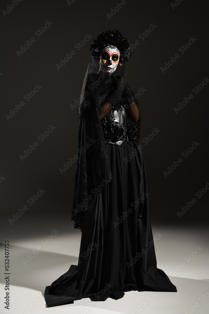 full length of woman in spooky costume and mexican day of dead makeup standing on black background.