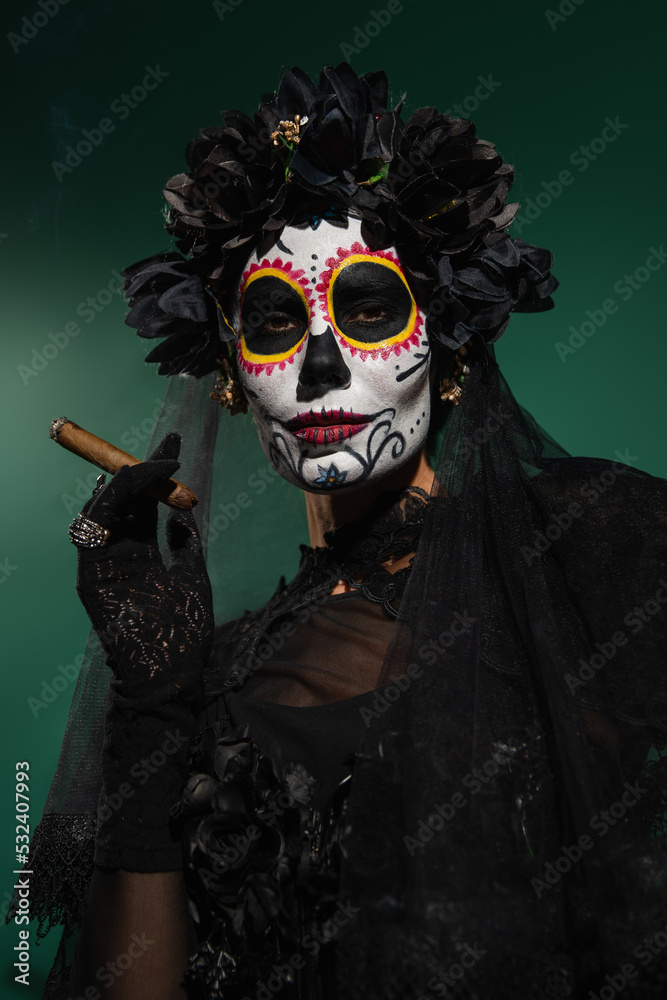 Woman in mexican day of death makeup and black costume holding cigar on green background.