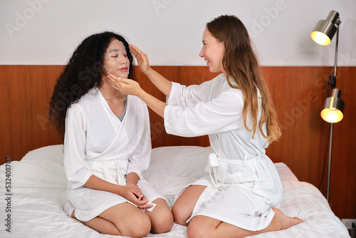 Young happy diversity ethnic beautiful woman in bathrobe sit on bed and doing cosmetic white facial mask for skin care and treatment. Women lesbian couple daily LGBTQ lifestyle and love concept.