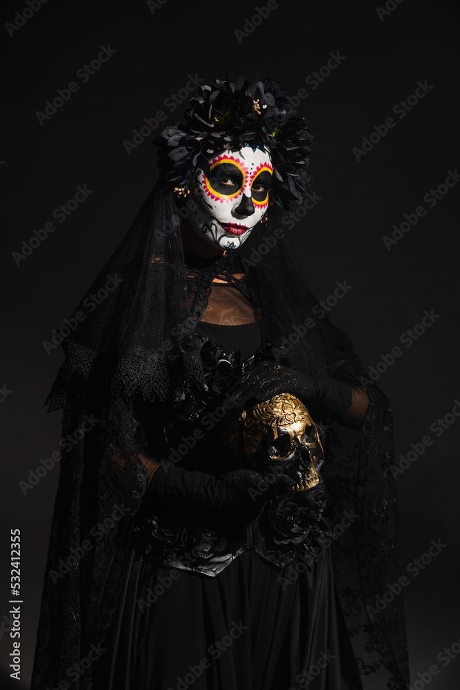 woman in creepy costume and catrina makeup holding golden skull isolated on black.