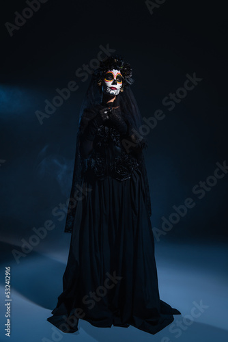 full length of woman in black witch dress and halloween makeup on dark background with blue light.