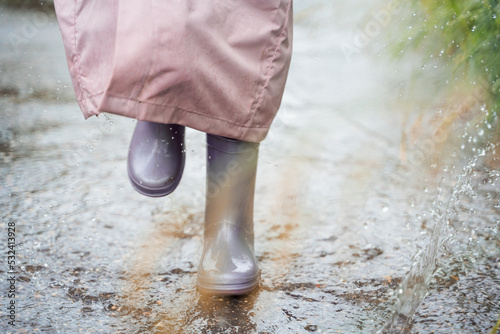 Little girl in pink waterproof raincoat  purple rubber boots funny jumps through puddles on street road in rainy day weather. Spring  autumn. Children s fun after rain. Outdoors recreation  activity