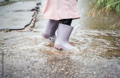 Little girl in pink waterproof raincoat, purple rubber boots funny jumps through puddles on street road in rainy day weather. Spring, autumn. Children's fun after rain. Outdoors recreation, activity © velirina