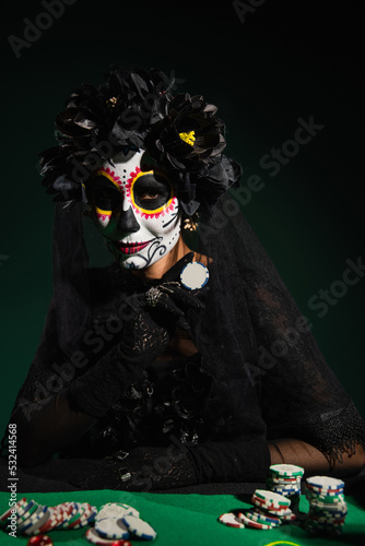 Woman in santa muerte halloween costume holding playing chip on dark green background.