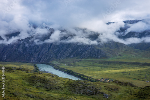 A large mountain range is covered with clouds early in the morning before sunrise. A beautiful landscape with a river in blue and green colors.