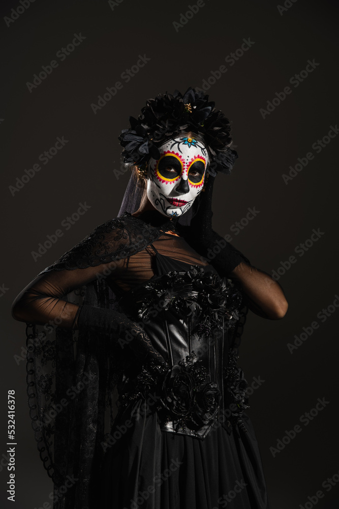 woman in dark halloween costume and sugar skull makeup posing with hand on waist isolated on black.