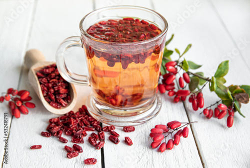 Berberis vulgaris also known as common barberry, European barberry or barberry tea drink in class mug in home kitchen. Dried and fresh berries for decoration on vintage white wood board background. photo