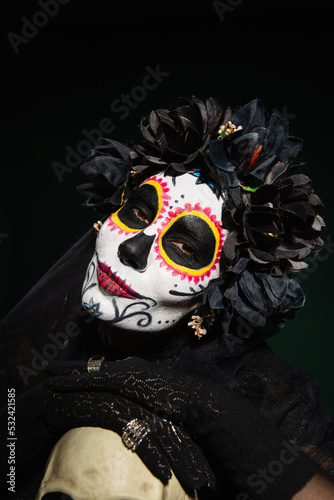 Woman in mexican santa muerte costume looking at camera near skull isolated on black.